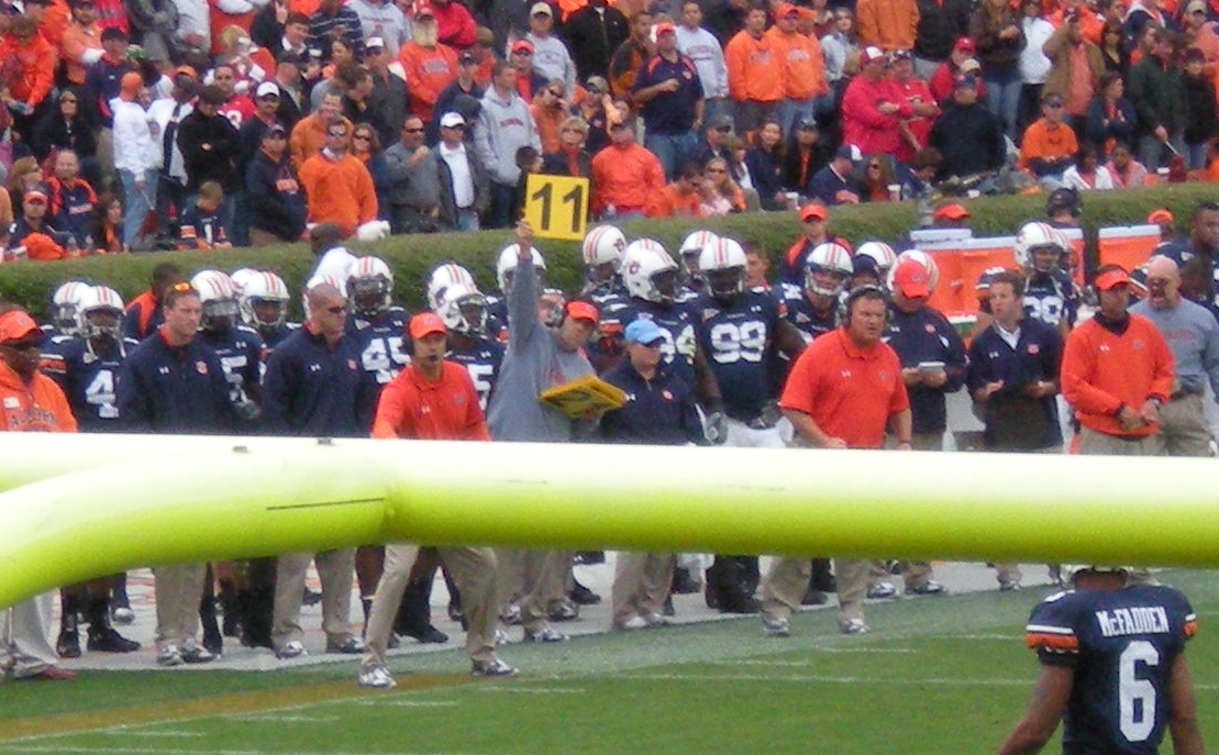Color photograph of a coach on the sideline of a game holding up a play sign. In this case, he has one hand raised over his head with a yellow sign reading '11' in black text. All around him are coaching staff and players on his team.