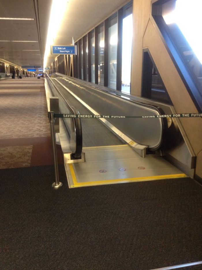 The photograph
depicts a long carpeted airport hallway with a moving walkway that
takes up the right half of the image. The walkway seems to still be
in operation, but it is blocked with a barricade labeled “Saving
Energy for the Future.”