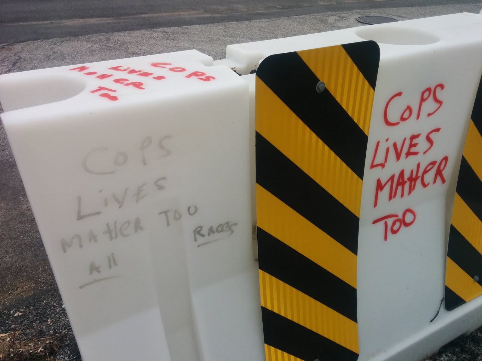 Photo of graffiti on a white road block. The letters are bold, scrawled in black and red permanent marker. The black letters, somewhat faded, read, 'Cops lives matter too' and underneath that, in underlined black text, 'all races.' On the other side of the road block and on top of it, this time written in red, the statement 'Cops lives matter' appears again.