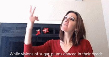 Figure 2 shows me, a young woman with shoulder-length brown hair, wearing a red long-sleeve shirt with stockings on a chimney behind me. I am signing VISIONS and looking upward while white captions at the bottom of the screen read “While visions of sugar-plums danced in their heads.”