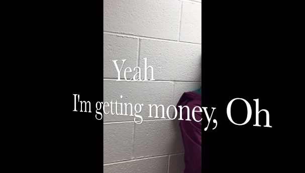 This screenshot shows a white wall and part of a woman's shoulder leaning on the wall. Large white captions appear diagonally on the screen and read “Yeah I'm getting money, Oh.”