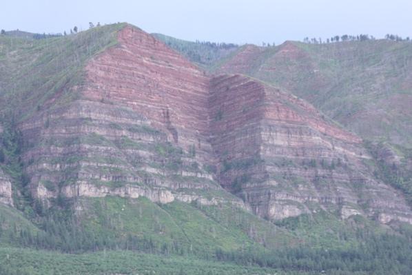 Photograph of a bluff cliff with layered red, grey, and beige sedimentary layers. Vegetation of oak brush and aspen and pine trees covers the top of the bluff, as well as the valley below.